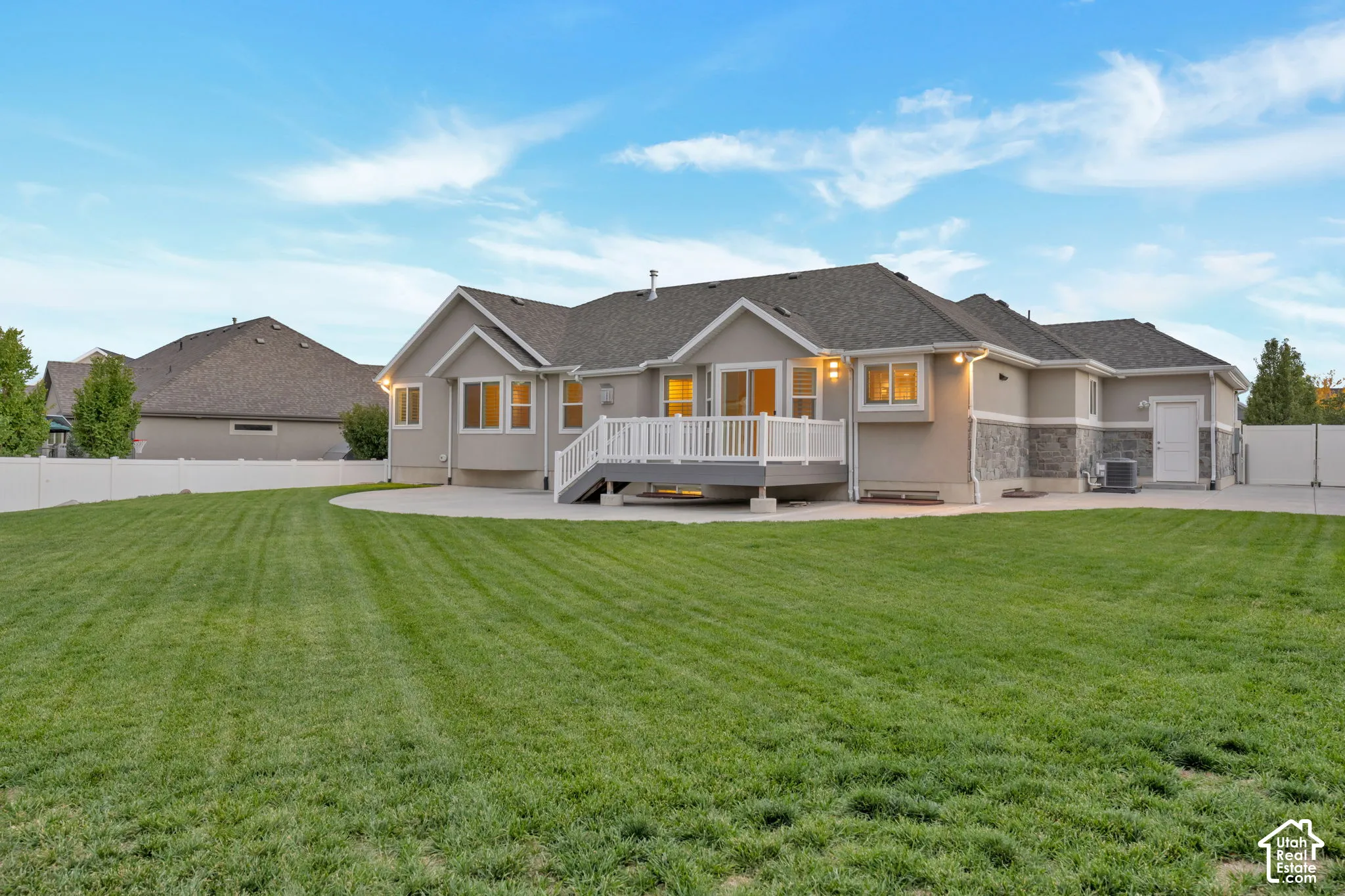 Rear exterior featuring a stone veneer foundation, expansive manicured lawn, concrete patio, elevated deck with white railing, and full vinyl privacy fencing