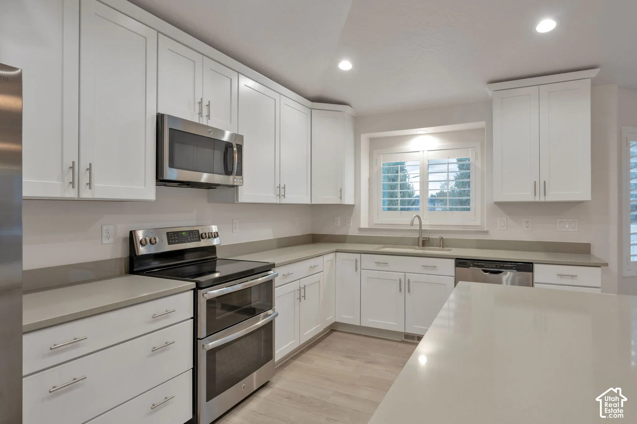 Kitchen featuring white shaker-style cabinetry, solid-surface countertops, stainless steel appliances, recessed ceiling lighting, and plantation shutters