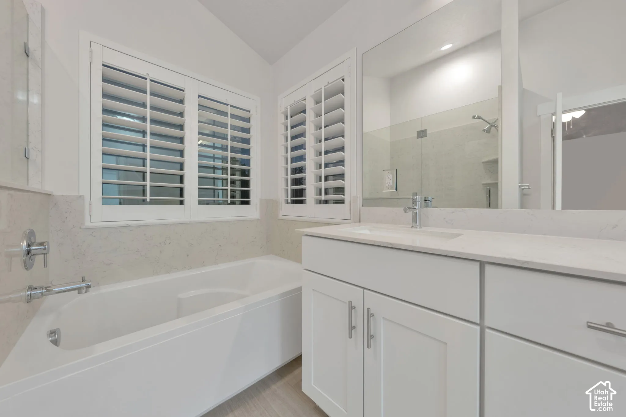 Bathroom featuring a drop-in soaking tub, solid surface countertop with an integrated sink, white shaker-style cabinetry, plantation shutters, and a wall-to-wall vanity mirror