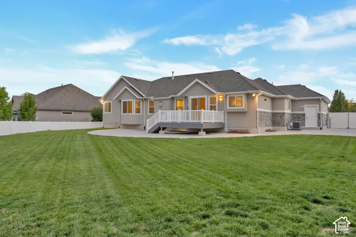 Rear exterior featuring a stone veneer foundation, expansive manicured lawn, concrete patio, elevated deck with white railing, and full vinyl privacy fencing