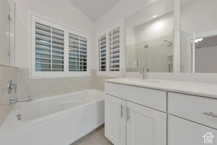 Bathroom featuring a drop-in soaking tub, solid surface countertop with an integrated sink, white shaker-style cabinetry, plantation shutters, and a wall-to-wall vanity mirror