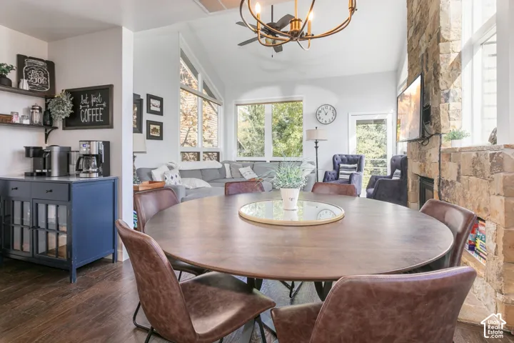 Vaulted ceiling with a chandelier and ceiling fan, stone fireplace with an integrated media display, built-in coffee bar with shelving, wood-finish flooring, and large windows