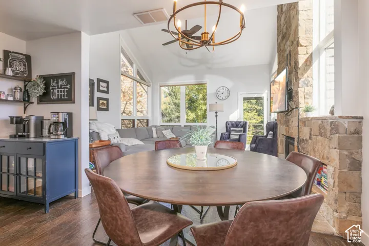 Open-concept living and dining area anchored by a round wood-finish dining table, featuring abundant natural light from multiple windows, a decorative brass chandelier, stone fireplace surround, and wood-finish flooring throughout