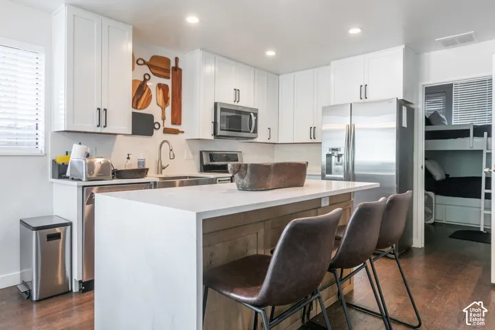 White shaker cabinetry with black hardware, stainless steel appliances, a center island with seating, a white countertop, and wood-finish flooring