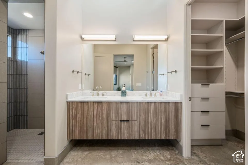 Floating vanity featuring a stone-look countertop with dual undermount sinks, wood-finish cabinetry, a large wall-mounted mirror with dual linear light fixtures, and a walk-in closet with built-in shelving and drawer storage