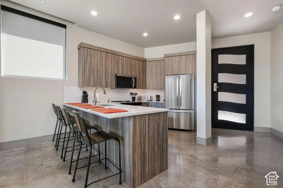 Kitchen featuring wood-finish cabinetry, stone-surface breakfast bar, stainless steel appliances, polished concrete flooring, and a contemporary entry door with frosted glass panel insets