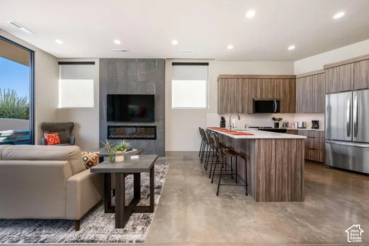 Open-concept space featuring polished concrete flooring, a floor-to-ceiling stone-clad fireplace, cabinetry with wood-finish detailing, and a kitchen island with stone-style countertops