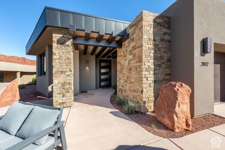 Contemporary exterior featuring stacked stone masonry, a dark metal roofline, exposed ceiling beams, a multi-panel glass entry door, and a concrete walkway