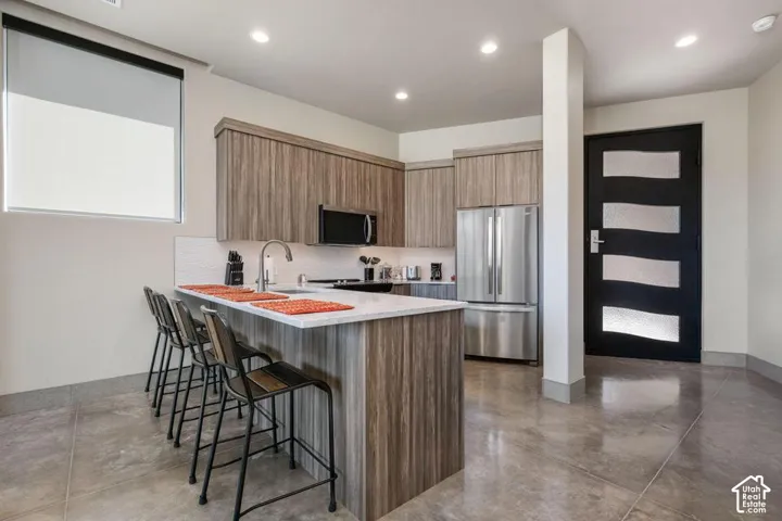 Kitchen featuring wood-finish cabinetry, stone-surface breakfast bar, stainless steel appliances, polished concrete flooring, and a contemporary entry door with frosted glass panel insets