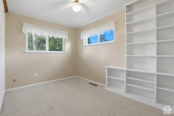 Built-in floor-to-ceiling shelving, carpeted flooring, two windows with horizontal blinds, and a central ceiling fan with integrated lighting