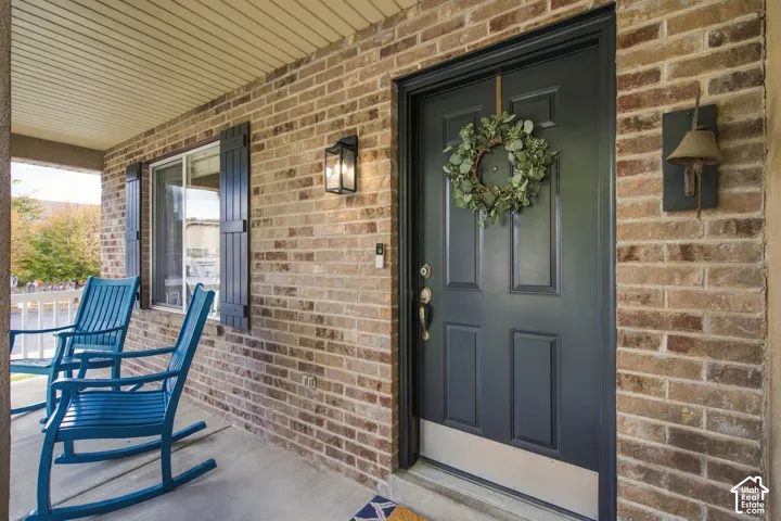 Covered front porch featuring a brick exterior facade, paneled door with a metallic kick plate, dark window shutters, and wall-mounted exterior lanterns