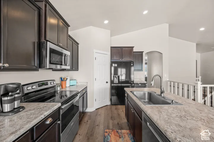 Kitchen featuring dark cabinetry, stone-finish countertops, stainless steel appliances, a center island with a drop-in sink, and wood-finish flooring