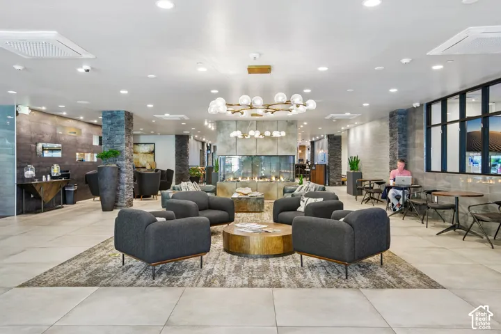 Open-concept lobby featuring large-format floor tiles, a central double-sided stone fireplace, recessed ceiling lighting, multi-arm spherical chandeliers, and textured stone accent pillars