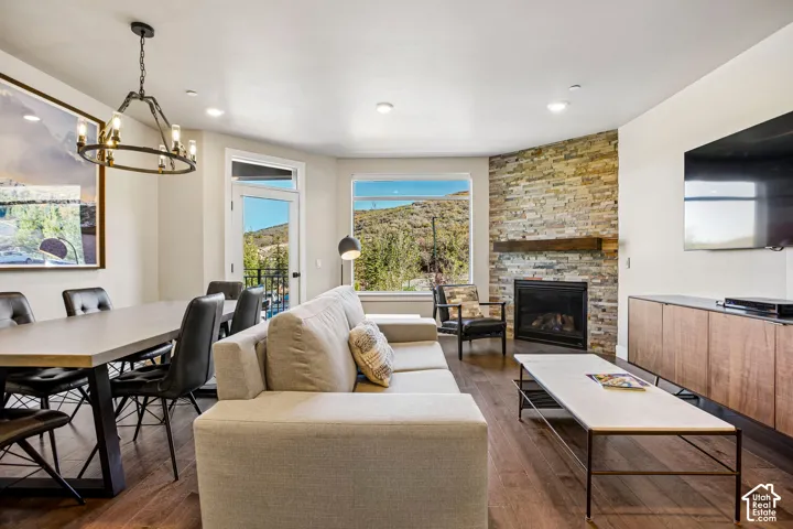 Living area featuring wood-finish flooring, a stone-accented fireplace with a timber mantel, a modern wagon-wheel chandelier, and large windows with mountain scenery