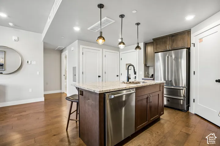 Kitchen featuring wood-finish flooring, a central island with a stone-composite countertop, stainless steel appliances, dark wood cabinetry, and industrial-style pendant lighting