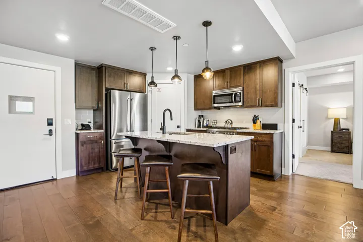 Kitchen featuring wood cabinetry, stone countertops, stainless steel appliances, a center island with a sink, and wood-finish flooring