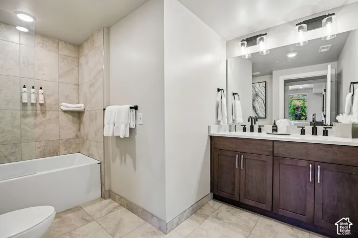 Bathroom featuring a dual-sink vanity with dark wood-finish cabinetry, a solid-surface countertop, and matte black fixtures, complemented by a tub-shower combination with floor-to-ceiling tile surrounds and stone-tile flooring