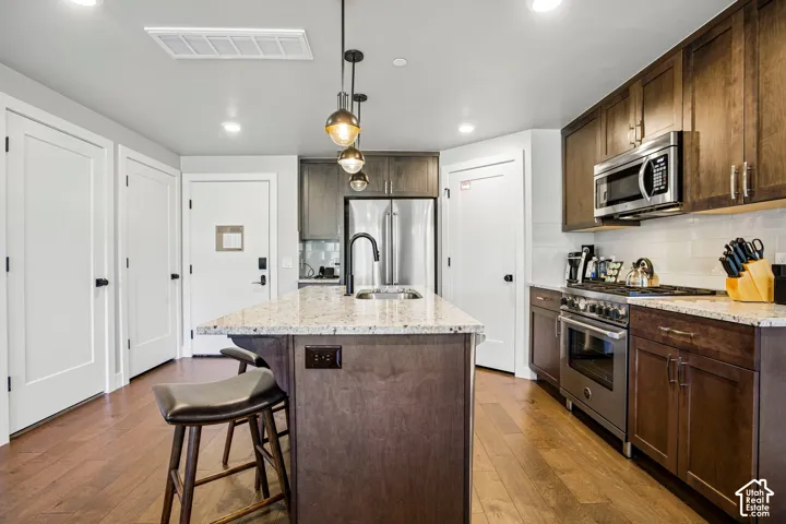 Kitchen featuring dark wood cabinetry, stone-surface countertops, stainless steel appliances, an island with an integrated sink, and wood-finish flooring