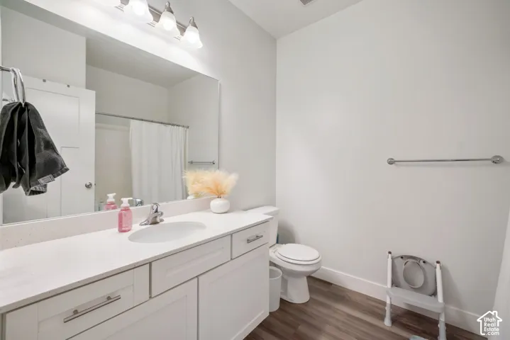 Bathroom vanity featuring a light-toned solid surface countertop, an undermount oval sink, shaker-style cabinetry with brushed metal hardware, a wall-to-wall vanity mirror, and wood-finish flooring