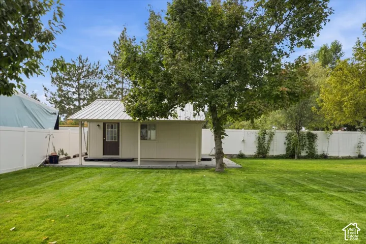 Backyard featuring a detached outbuilding with a metal roof, concrete patio, white vinyl privacy fencing, and mature trees