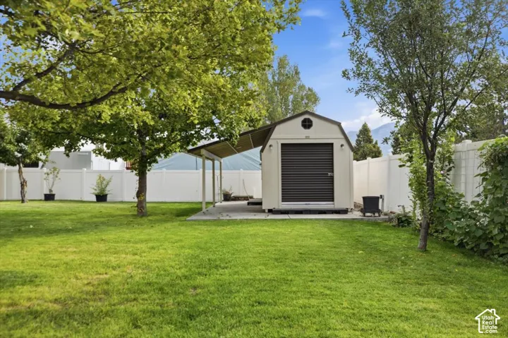 Backyard featuring a grass lawn, white vinyl fencing, mature trees, and a detached storage structure with an attached metal-roofed carport and concrete pad
