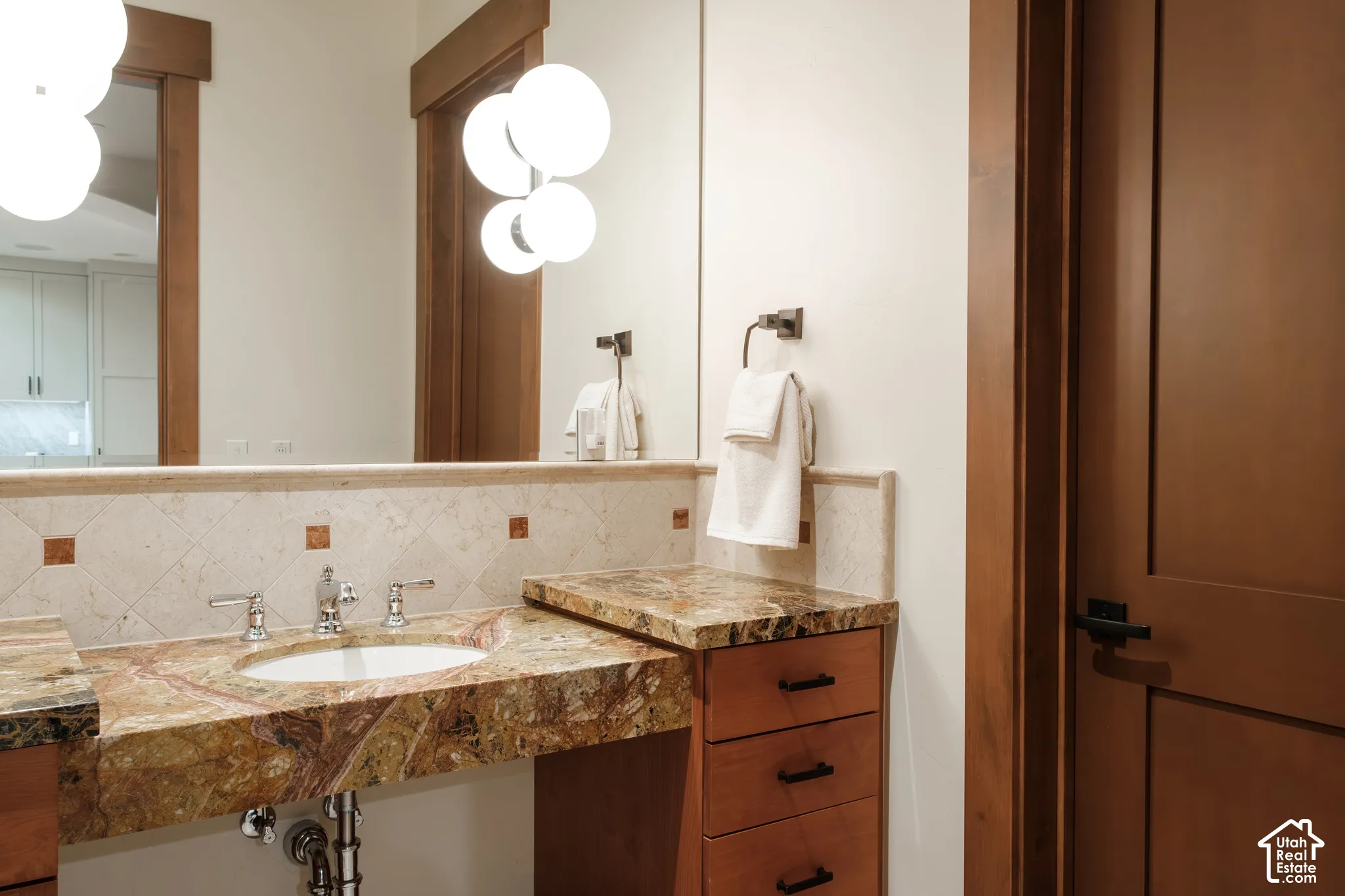 Vanity featuring natural stone countertop, integrated basin, diamond-patterned tile backsplash with decorative stone insets, wood cabinetry with matte black hardware, and multi-globe wall-mounted light fixtures