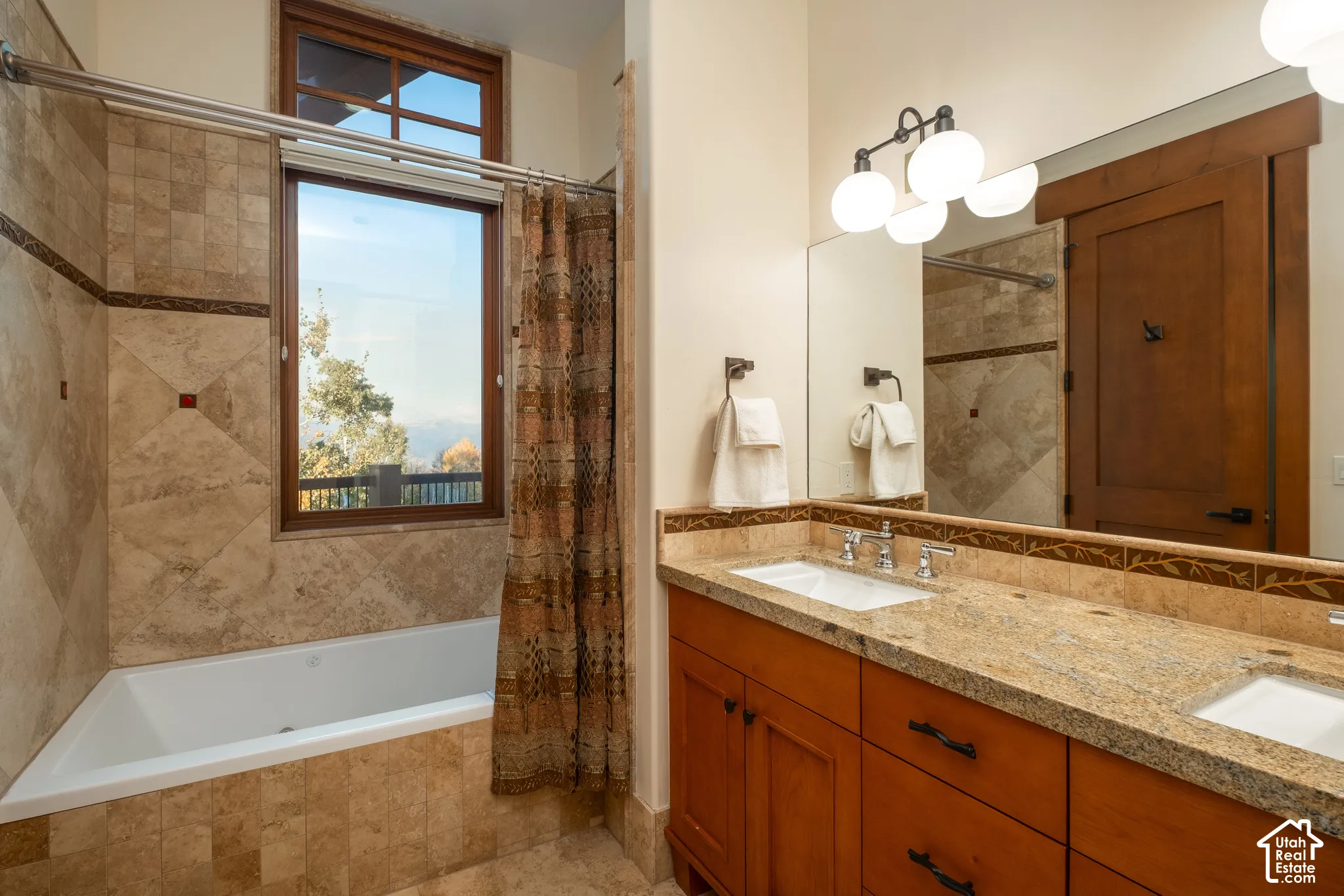 Bathroom featuring a dual-sink vanity with stone countertops, tiled bathtub surround with an inset decorative border, wood-framed cabinetry, and a large window