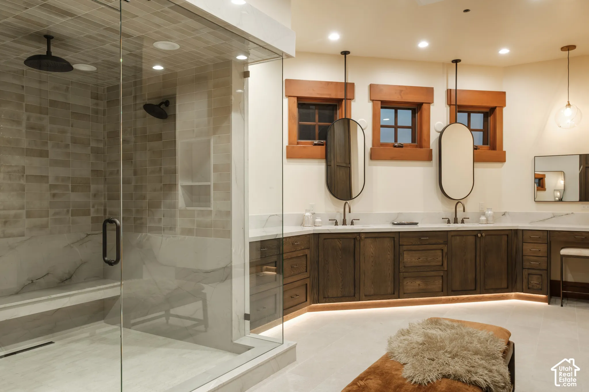 Walk-in glass-enclosed shower with a stone bench and rainfall showerhead, dual-sink vanity with stone countertop, wood-finish cabinetry, and oval wall-mounted mirrors