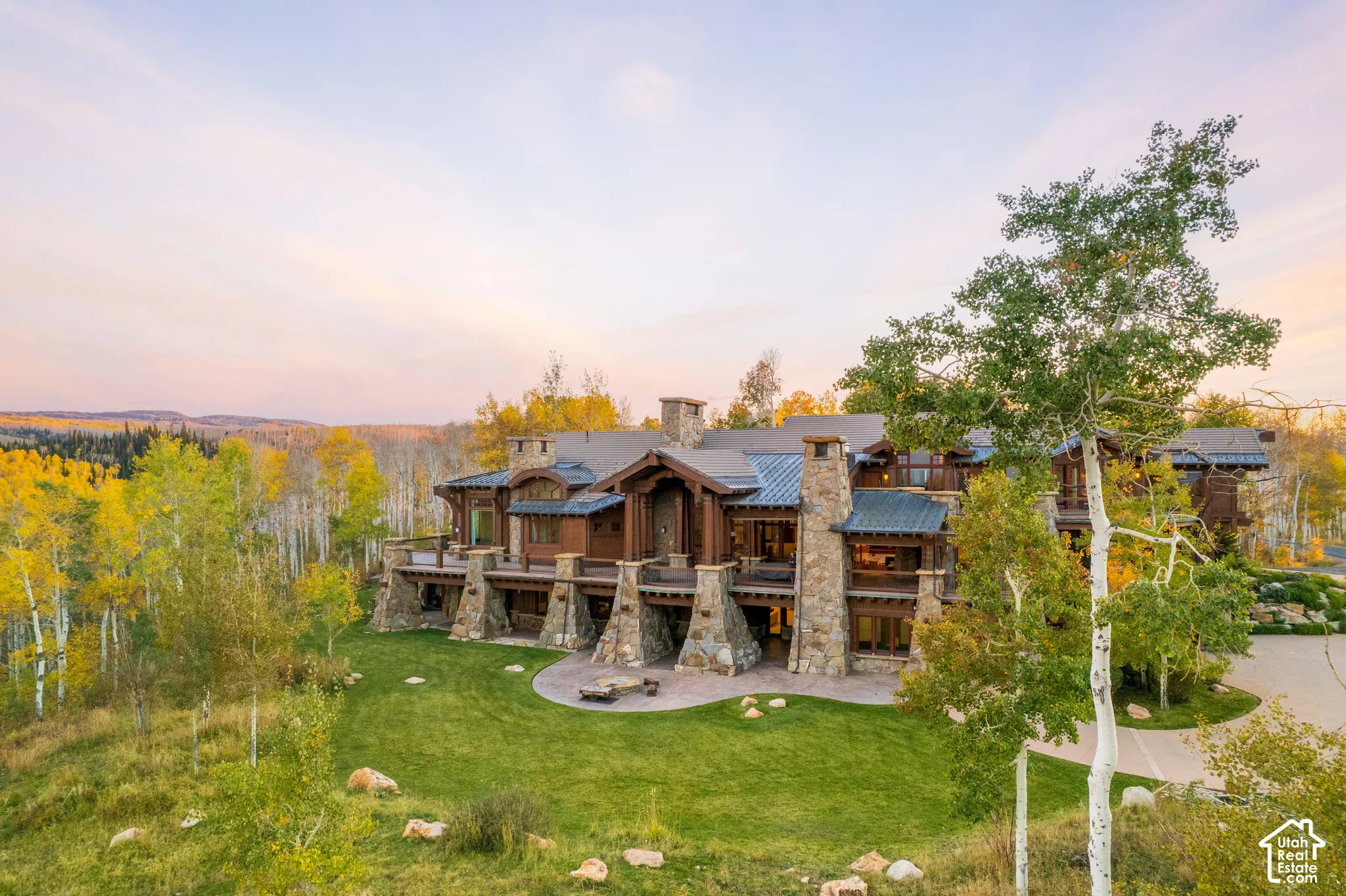 Mountain-style exterior featuring stone masonry, exposed timber framing, metal roofing, expansive wraparound decks, and a paved stone patio