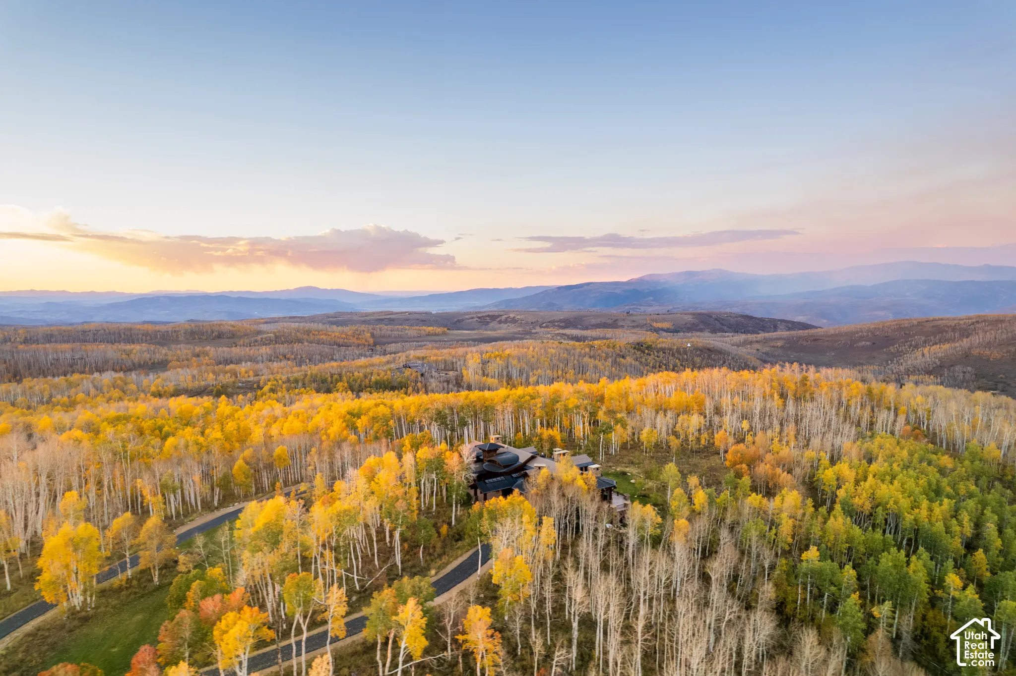 Aerial perspective featuring a multi-level structure nestled within a dense forest, surrounded by mature deciduous trees and a private paved driveway, set against a backdrop of mountain ranges