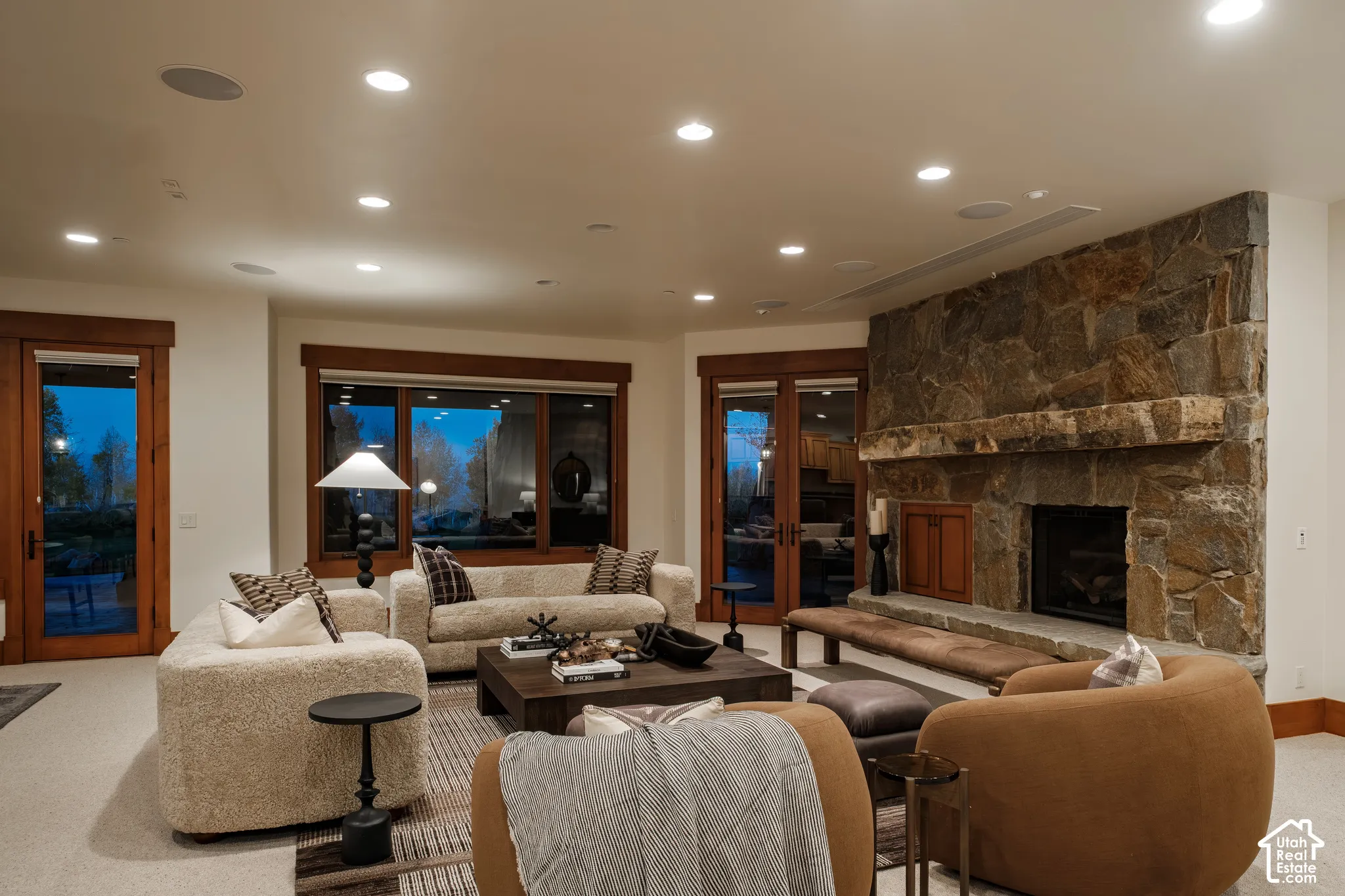 Floor-to-ceiling stone fireplace with a thick timber mantel and raised stone hearth, natural wood window and door casings, and recessed ceiling lighting