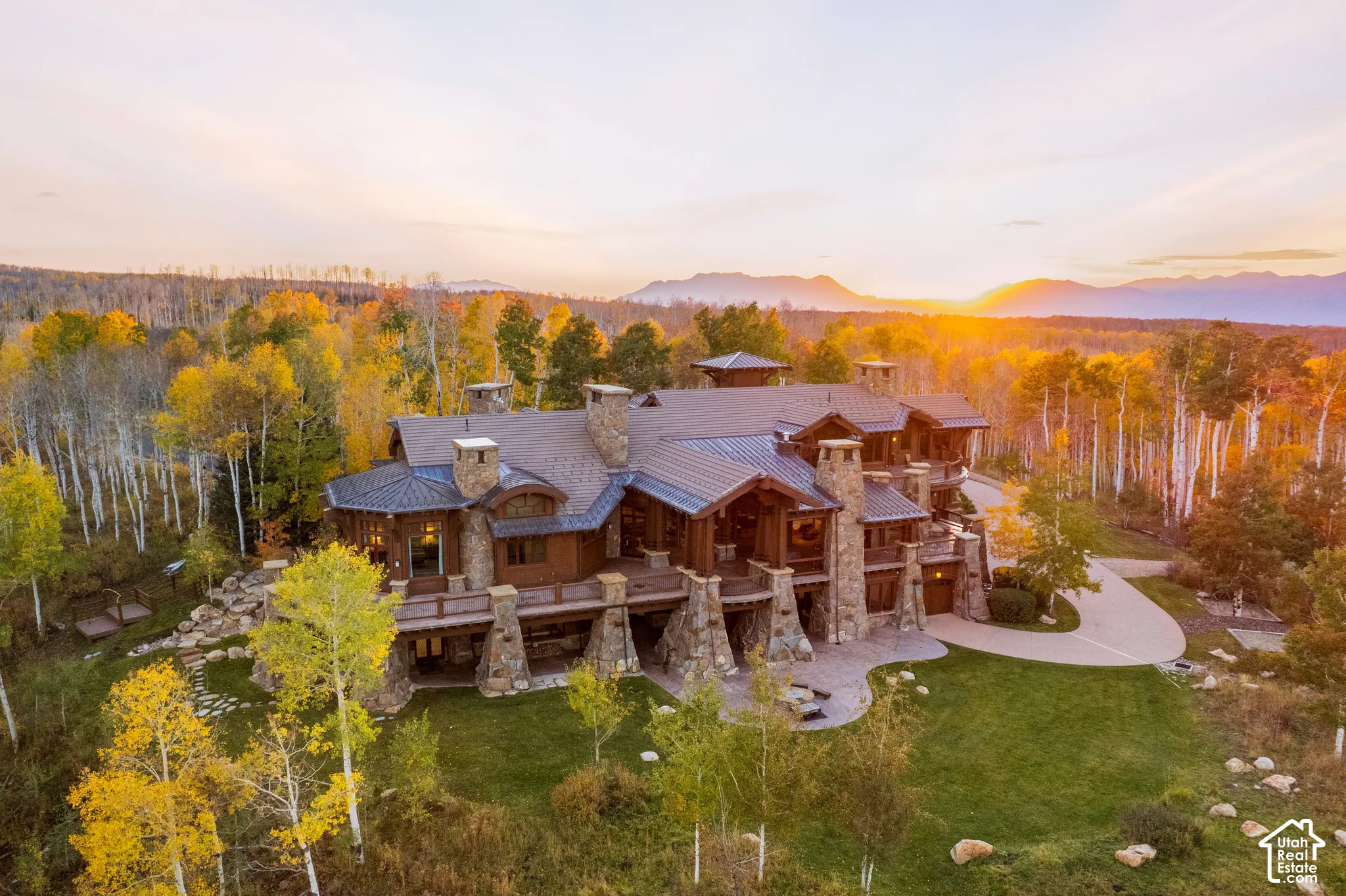 Custom mountain estate featuring stone masonry exterior, metal roof, wrap-around timber deck, stone chimney stacks, and concrete driveway