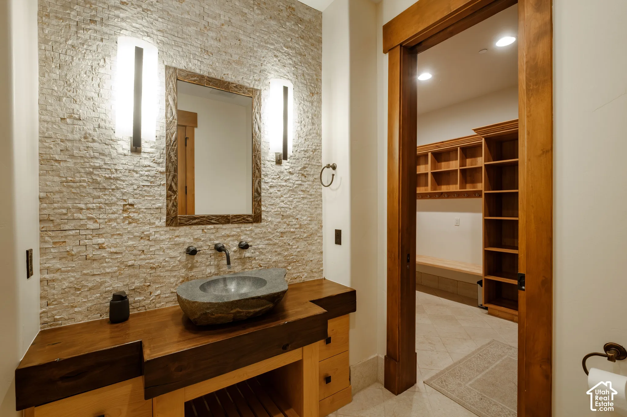Stacked stone accent wall with dual wall-mounted sconces, custom wood vanity featuring a natural stone vessel sink, and built-in wood shelving units