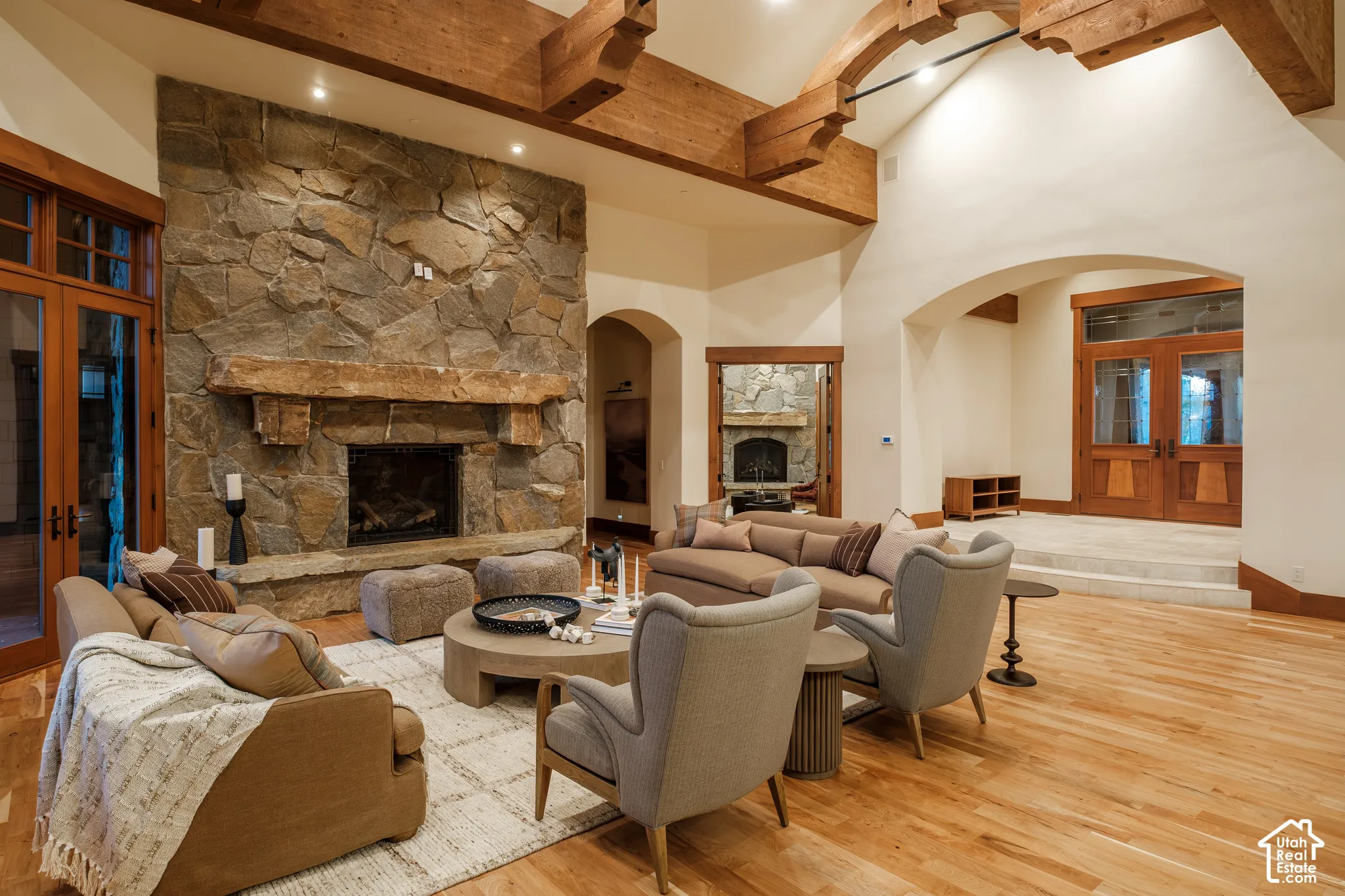 Vaulted ceiling with exposed wood beams, full-height stone fireplace, substantial wood mantel, arched entryways, and wood-finish flooring