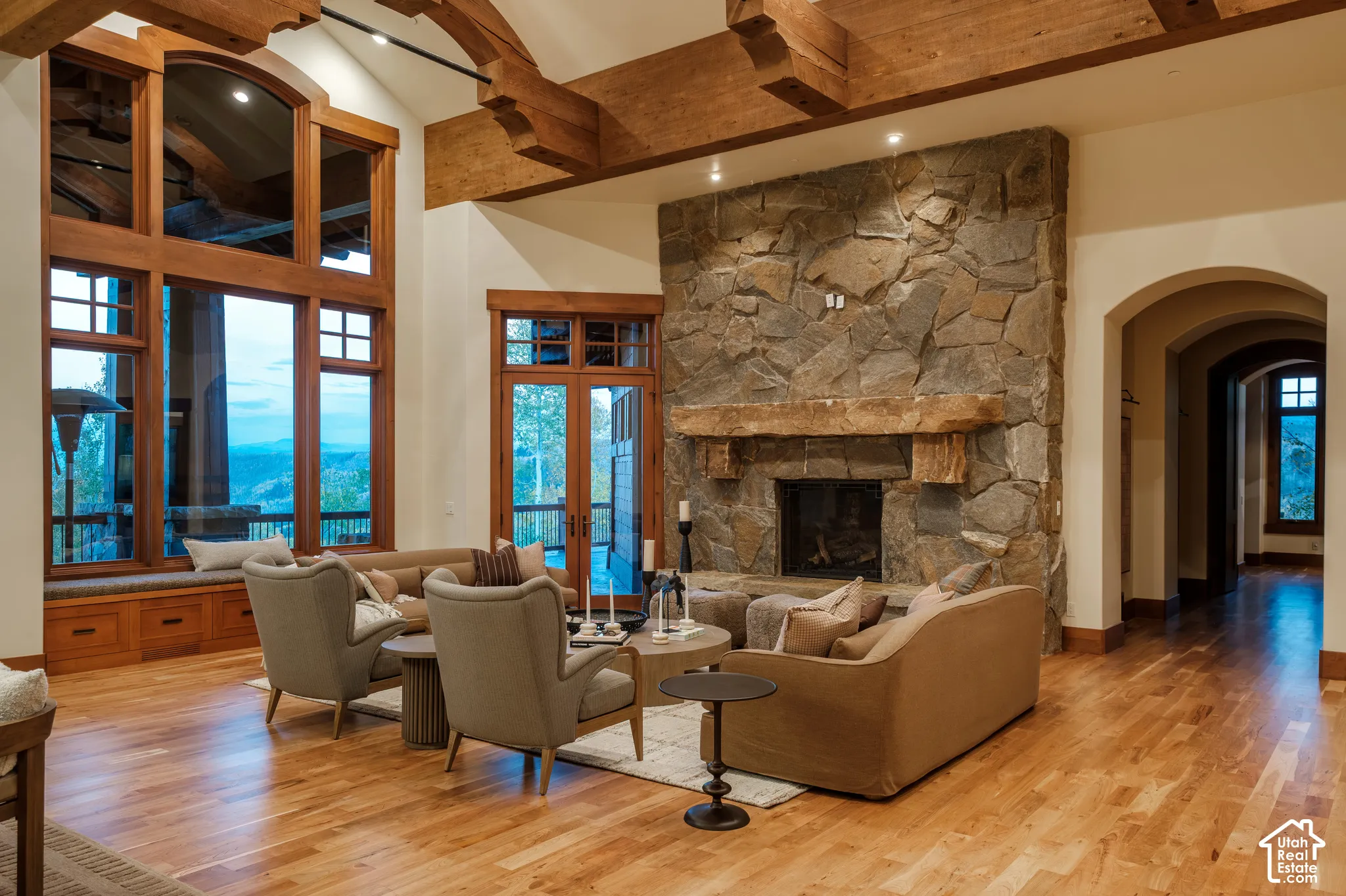 Vaulted ceiling with exposed timber beams, floor-to-ceiling stone fireplace with a thick timber mantel, wood-finish flooring, and expansive windows with custom wood trim