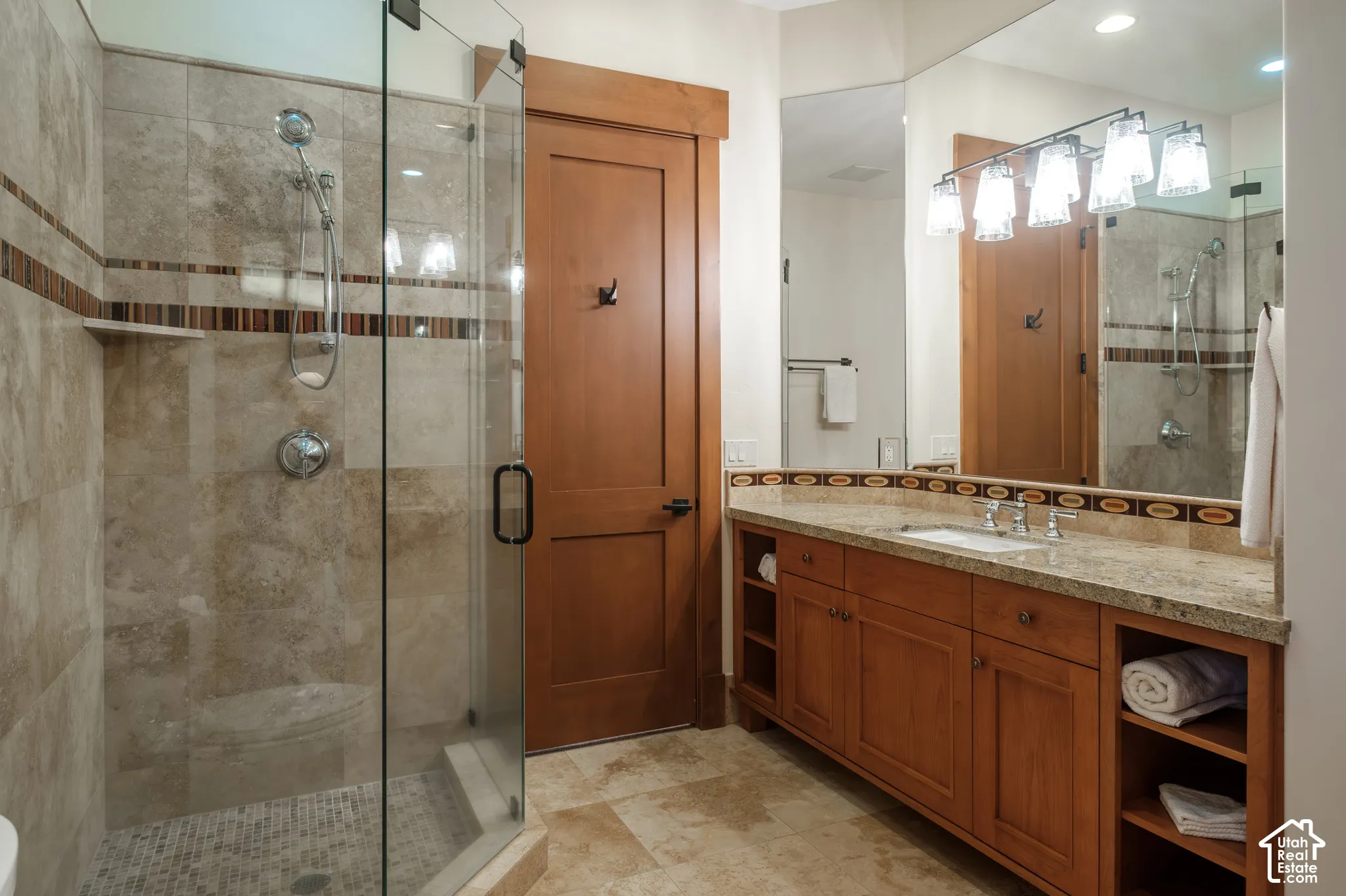 Bathroom featuring a glass-enclosed shower with stone tile surround, stone slab vanity countertop with an integrated rectangular sink, natural wood cabinetry with open side shelving, and a large wall-mounted mirror with multi-light fixture