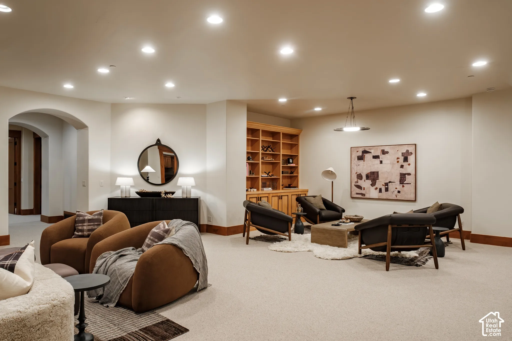 Expansive basement recreation area featuring recessed ceiling lighting, wood-finish built-in cabinetry with display shelving, wood-frame seating elements, and plush carpeting throughout
