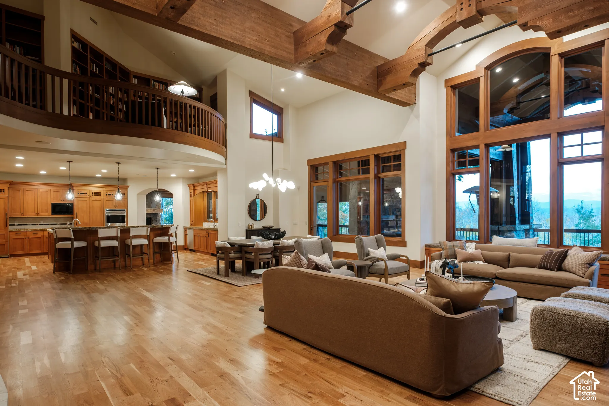 Expansive great room featuring vaulted ceilings with exposed timber trusses, wood-finish flooring, floor-to-ceiling multi-pane windows, and a wrap-around wooden mezzanine balcony with built-in cabinetry