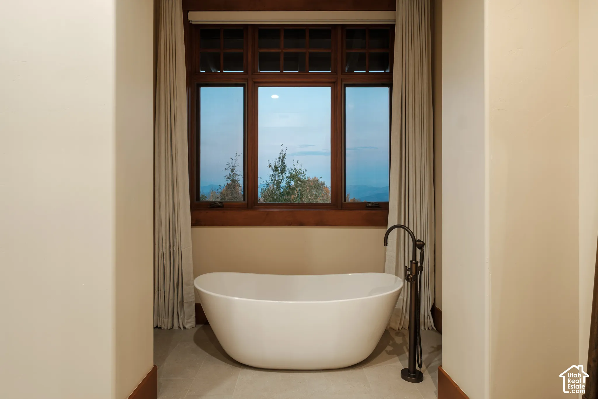 Freestanding soaking tub with a floor-mounted dark metal faucet, positioned before a multi-pane window with dark wood trim and mountain scenery