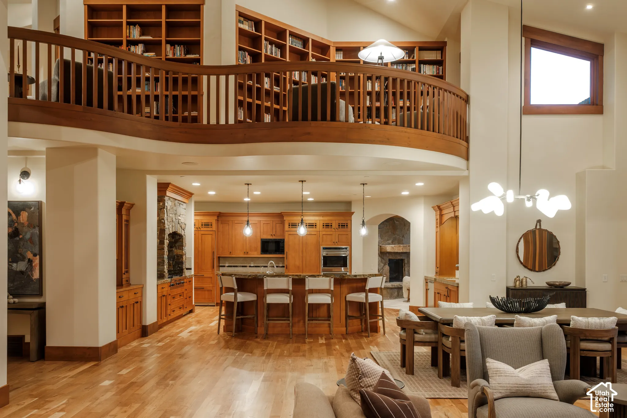 Expansive open-concept interior featuring wood-finish flooring, a curved wood-railing mezzanine, floor-to-ceiling custom built-in bookshelves, and a kitchen with wood cabinetry and stone countertops