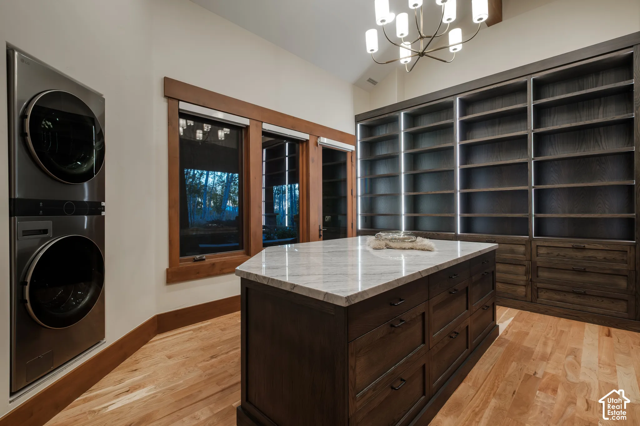 Multi-functional room featuring floor-to-ceiling built-in shelving with integrated LED lighting, a center island with a stone countertop, wood-finish flooring, and a stacked stainless steel washer and dryer unit