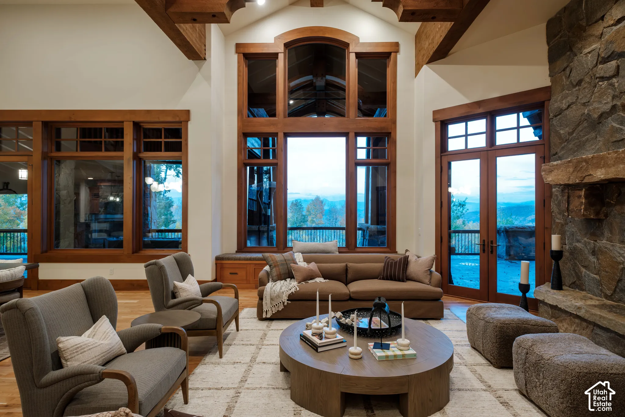 Vaulted ceiling featuring exposed timber beams, full-height windows with wood trim, floor-to-ceiling natural stone fireplace, and wood-finish flooring