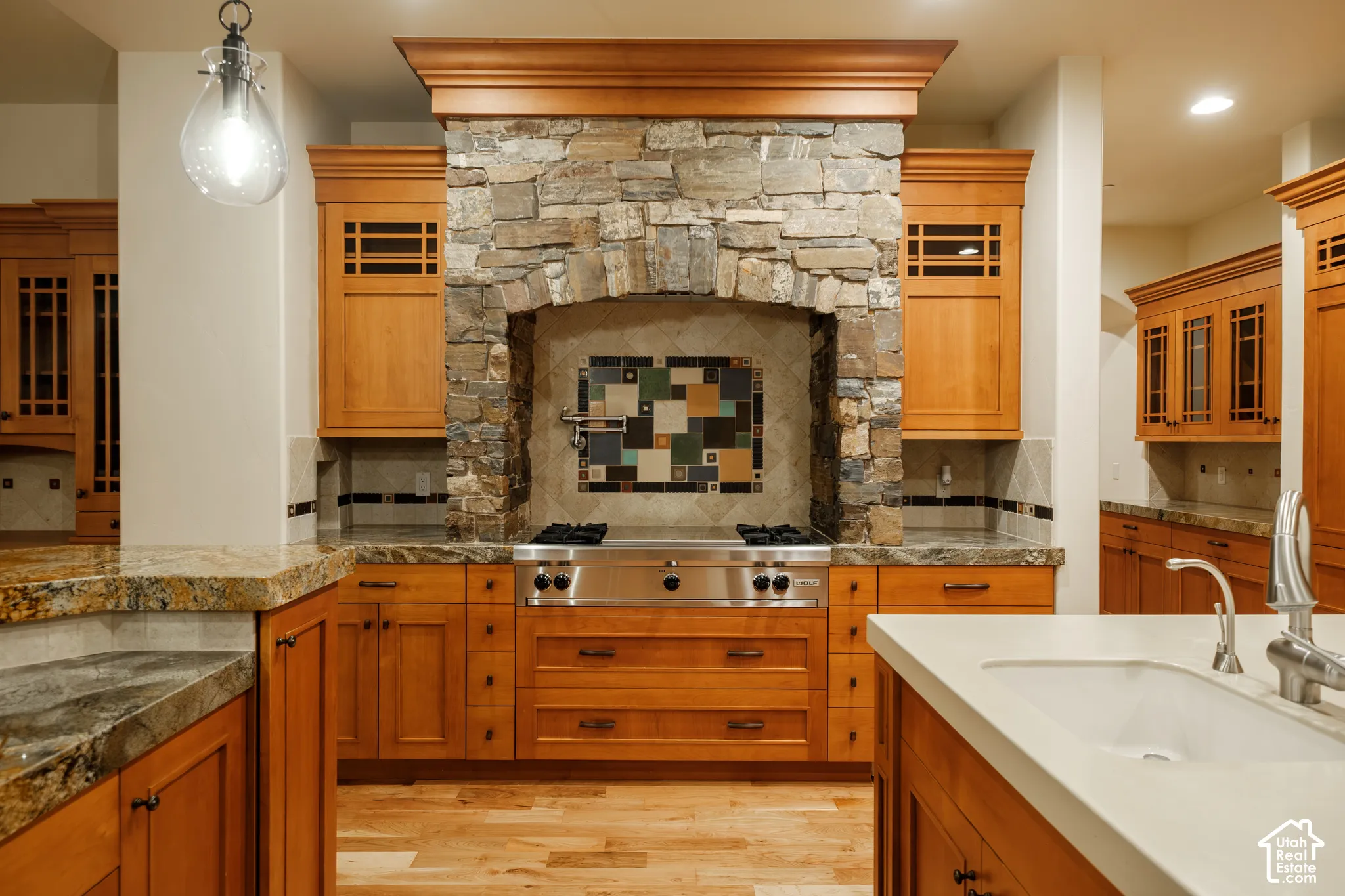 Custom cabinetry with glass-panel inserts, stone-surround cooktop alcove with decorative tile backsplash, professional-grade stainless steel gas range, stone countertops, and wood-finish flooring
