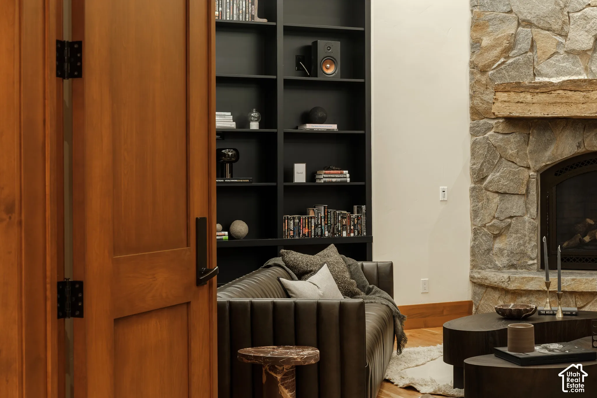 Floor-to-ceiling built-in matte black shelving, full-height natural stone fireplace surround, heavy timber mantel, and solid wood-finish door with black hardware