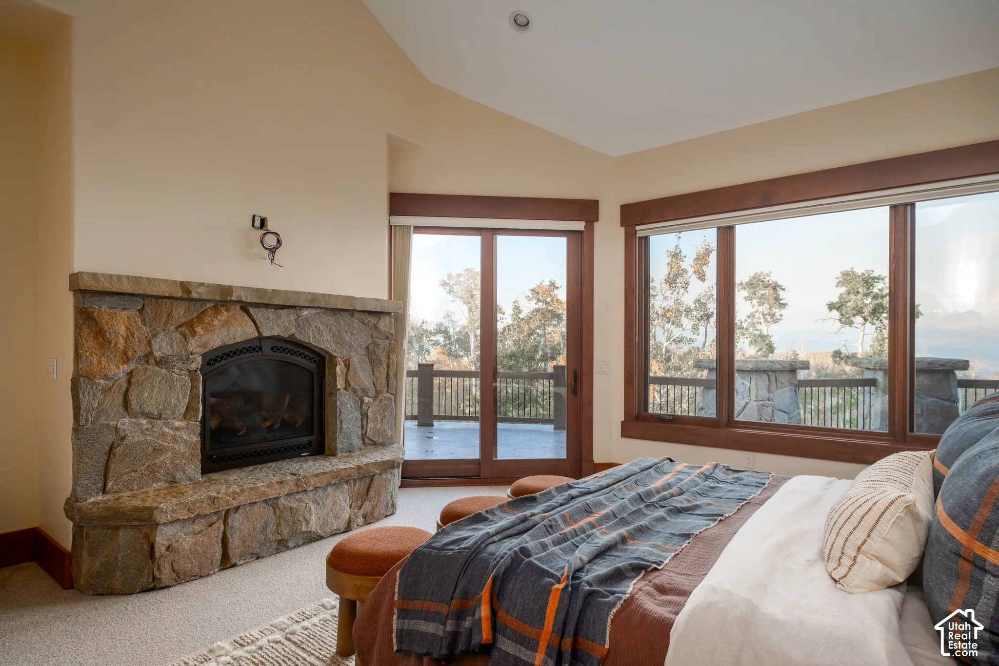 Full-height stacked-stone fireplace with a natural stone hearth, vaulted ceiling, large picture windows with wood trim, and sliding glass doors providing access to the deck