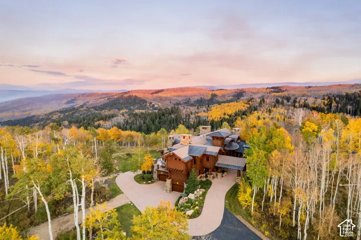 Mountain estate featuring a multi-gabled wood and stone exterior, metal roof, paved circular driveway, and stone masonry chimneys, set against a backdrop of mountainous terrain