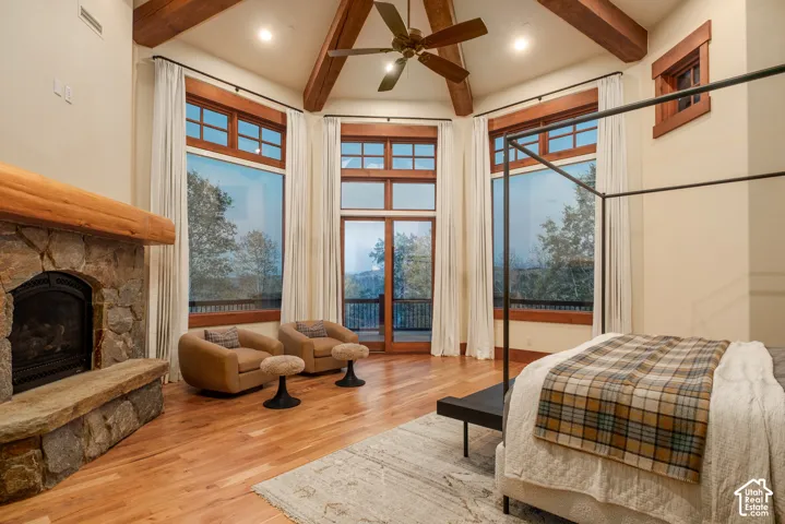 Vaulted ceiling with exposed wood beams, stone fireplace with a thick timber mantel, wood-finish flooring, floor-to-ceiling windows with wood trim, and a ceiling fan