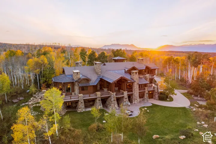 Custom mountain estate featuring stone masonry exterior, metal roof, wrap-around timber deck, stone chimney stacks, and concrete driveway