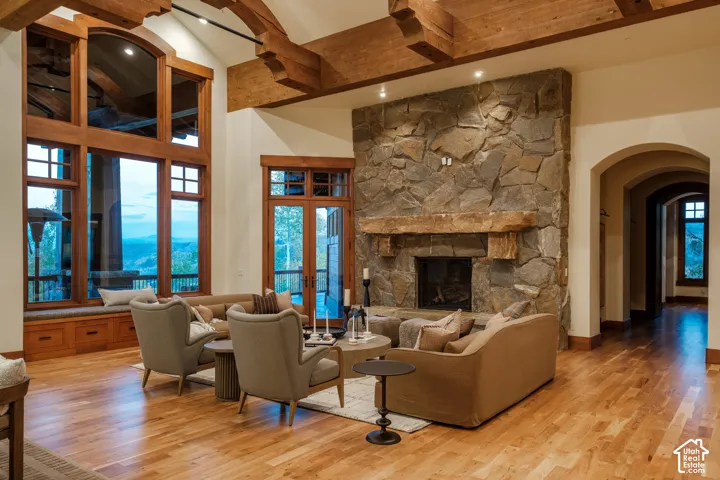 Vaulted ceiling with exposed timber beams, floor-to-ceiling stone fireplace with a thick timber mantel, wood-finish flooring, and expansive windows with custom wood trim