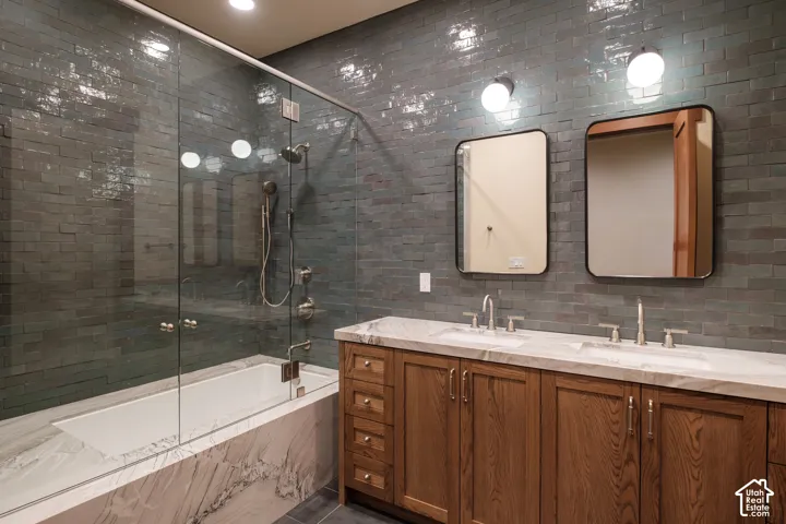 Custom wood-finish vanity with stone countertop, dual undermount sinks, glazed subway tile wall, and glass-enclosed bathtub with integrated shower hardware